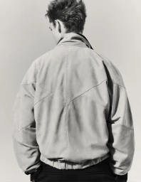 Back view of a male model wearing an oversized light-toned suede bomber jacket with geometric panelling against a plain backdrop.