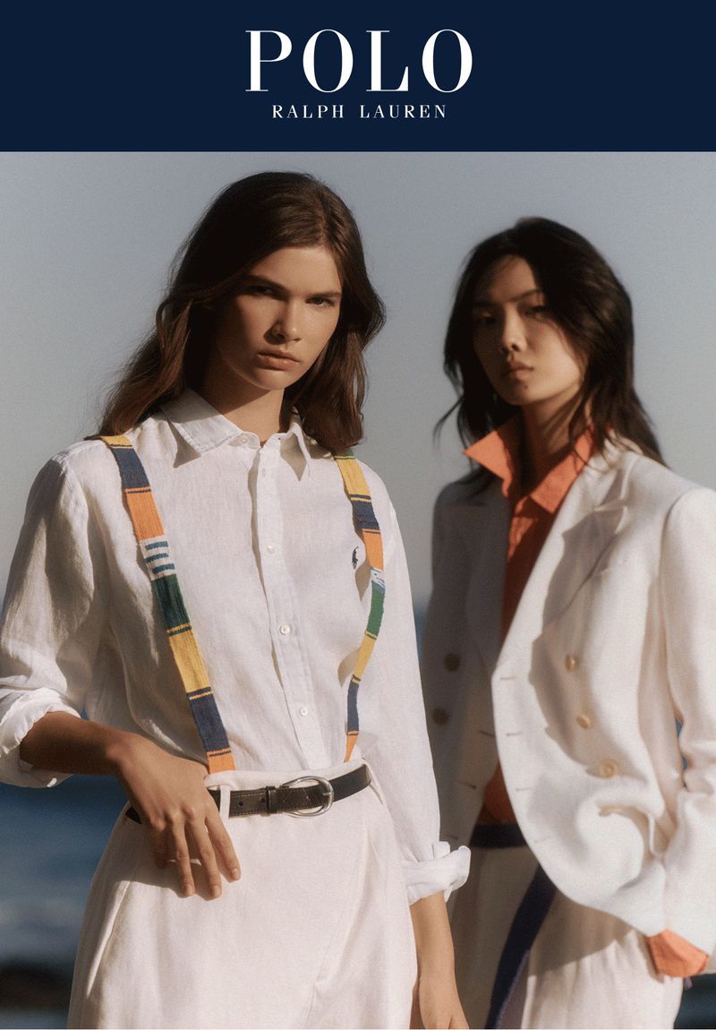 Two young women pose outdoors wearing white linen outfits, one with suspenders, for a Polo Ralph Lauren fashion shot.