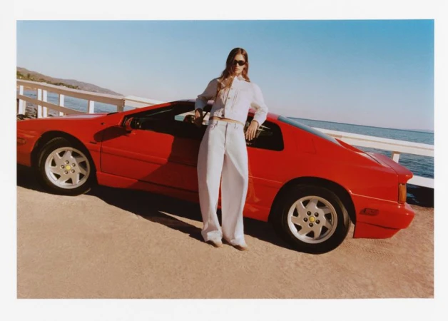 A female model in white denim poses near a red vintage sports car by the ocean, under bright sunlight showcasing fashion and automotive design.
