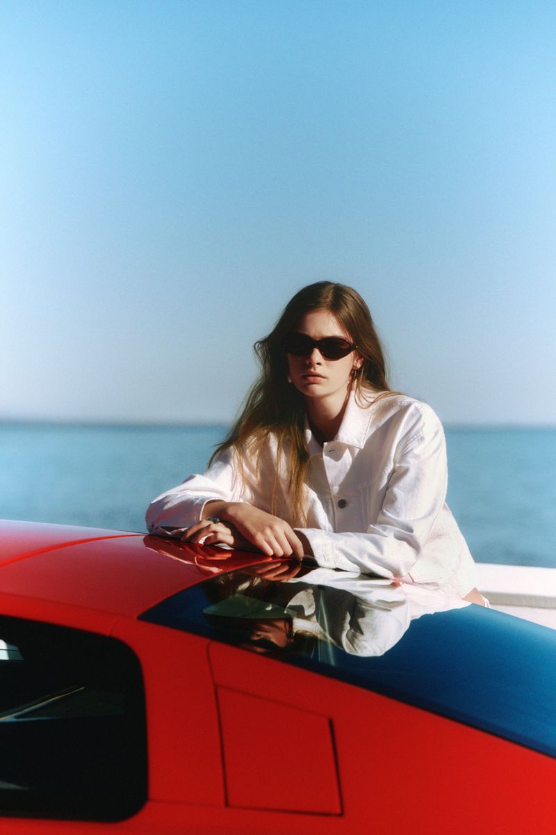 A woman with long hair and sunglasses poses on the hood of a red sports car against a blue sky and ocean backdrop on a sunny day.