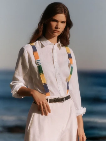 Fashion shot of a woman wearing a white collared shirt and suspenders, standing outside against an ocean backdrop, sunny and bright.