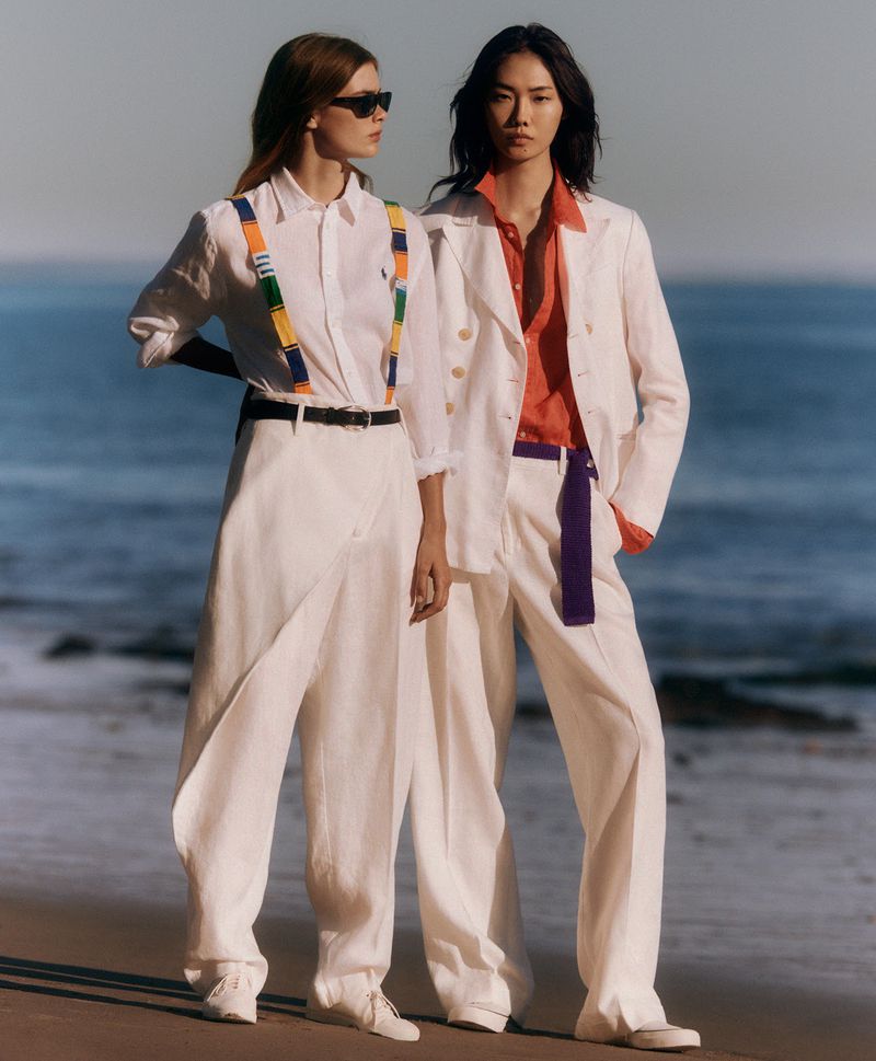 Two fashion models pose on a sandy beach in white linen outfits, featuring open shirts, wide-leg pants, and sunglasses, bathed in warm sunlight.