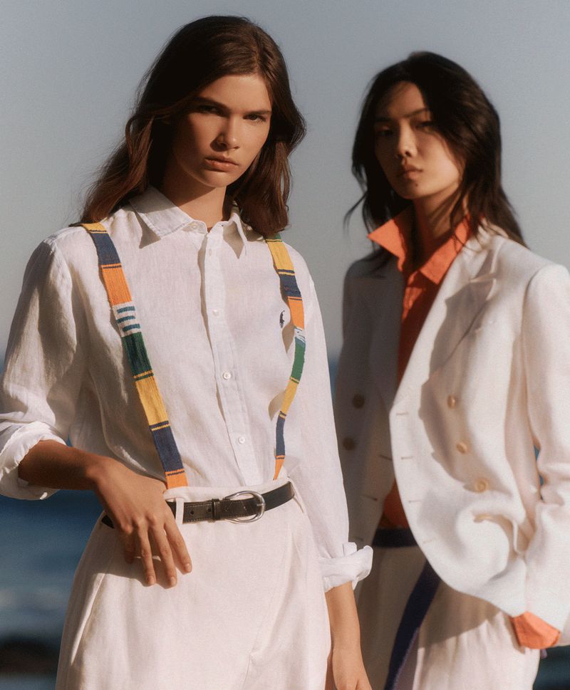 Two fashion models, one wearing suspenders, pose outdoors in sunlight wearing white linen clothing, against a blurred sky background.