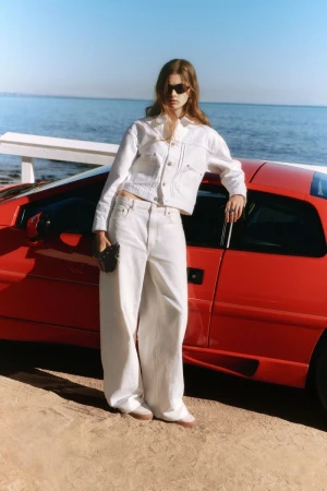 Model wearing a white denim jacket and matching wide-leg trousers leaning against a red sports car by the ocean.