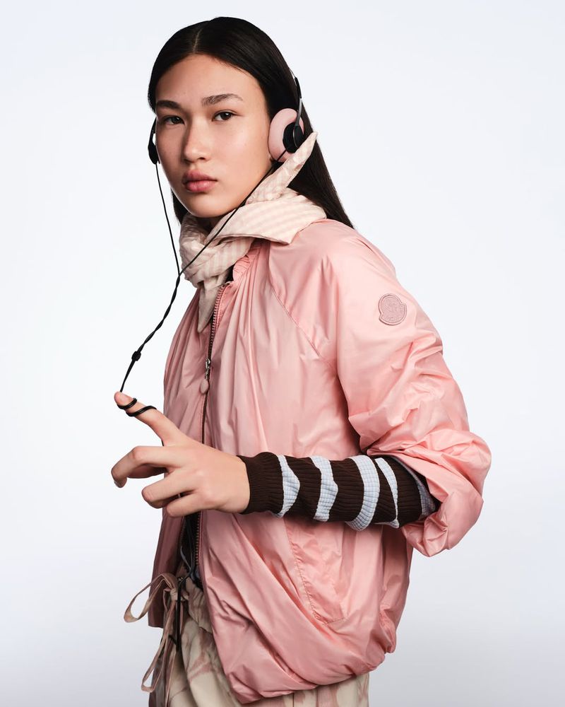 Fashionable young woman with long dark hair wearing pink jacket, headphones, and striped scarf in a bright, neutral indoor space.