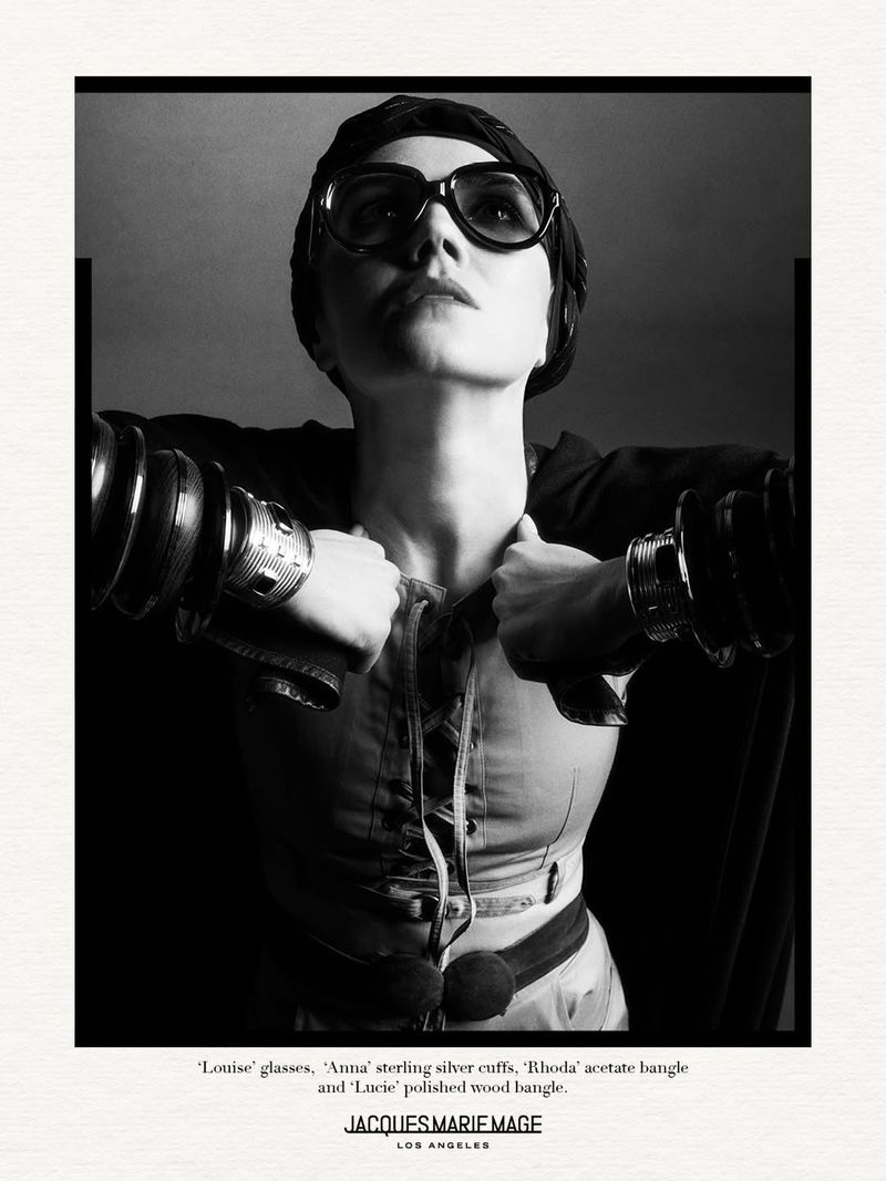 A woman poses in a black and white studio portrait wearing glasses, several bracelets, and a dark jacket.