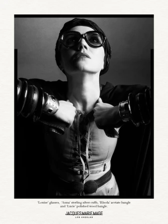 A woman poses in a black and white studio portrait wearing glasses, several bracelets, and a dark jacket.