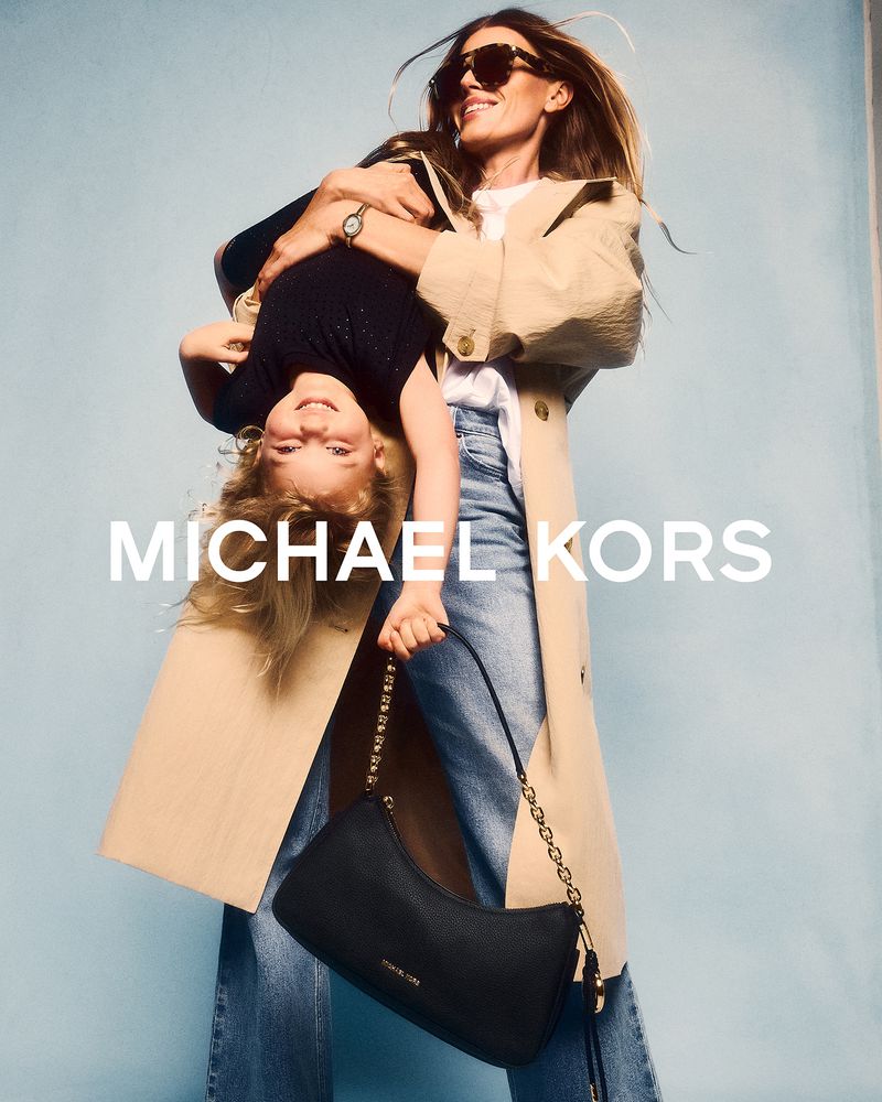 A joyful mother holds her daughter upside down, showcasing a stylish black Michael Kors handbag against a soft blue backdrop.