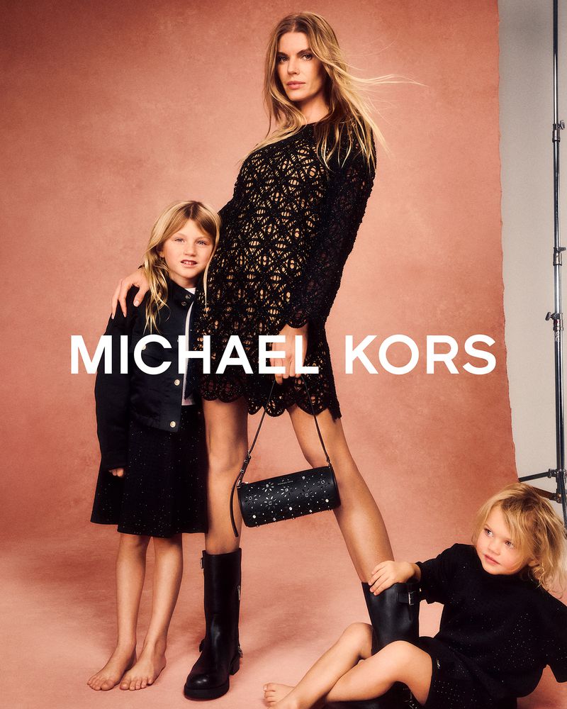 Three females pose in front of a blush-colored backdrop. The adult wears a black dress, holding a purse, while the children wear black dresses.