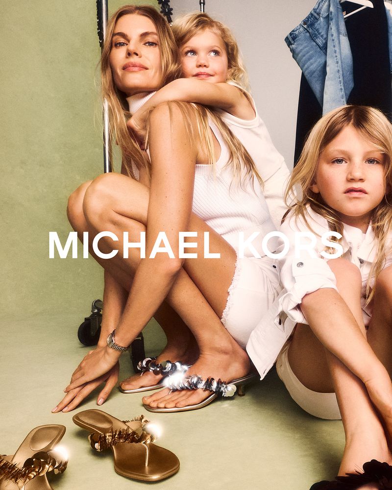 Fashion ad with a seated adult female, two children, and sandals; Michael Kors branding is visible, set against a neutral backdrop.
