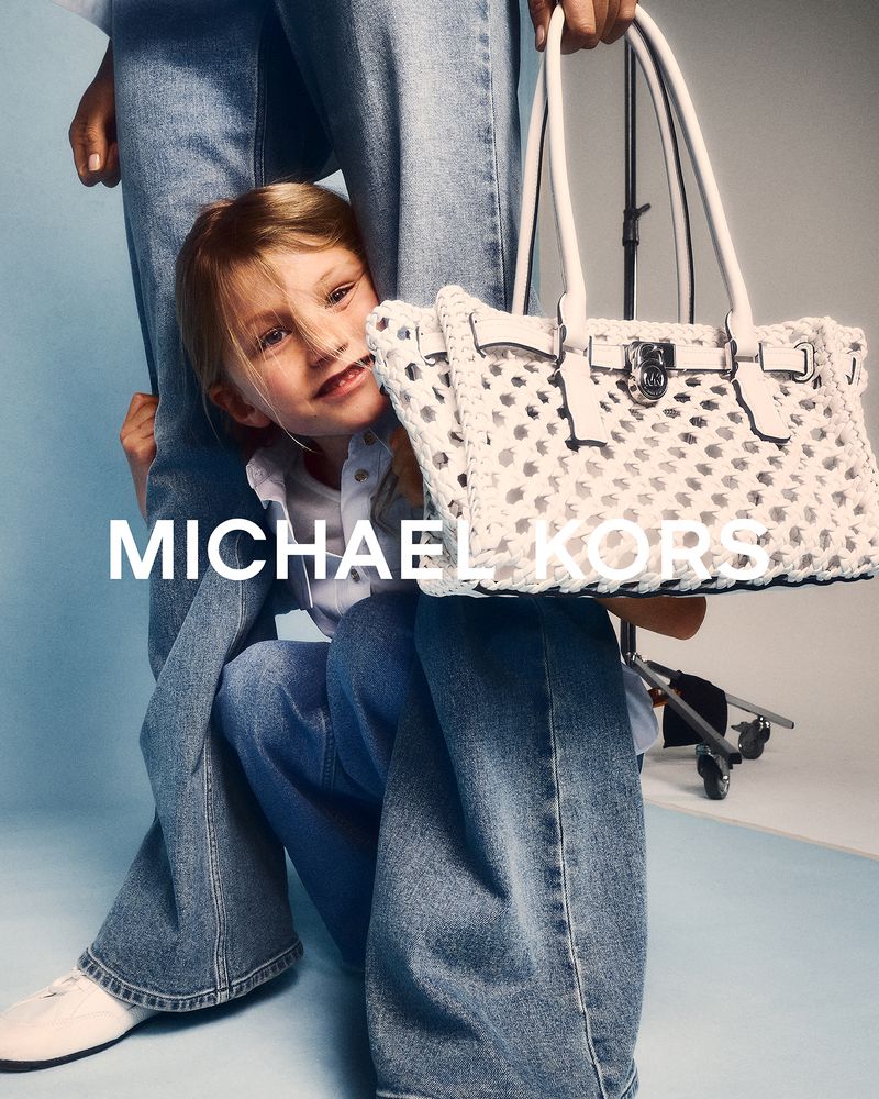 A child peeks out from behind an adult wearing jeans, holding a white woven handbag, against a bright studio backdrop.