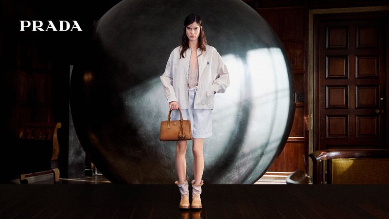 Adult female fashion model wears a light jacket and shorts while holding a tan purse in a studio with a sphere background.