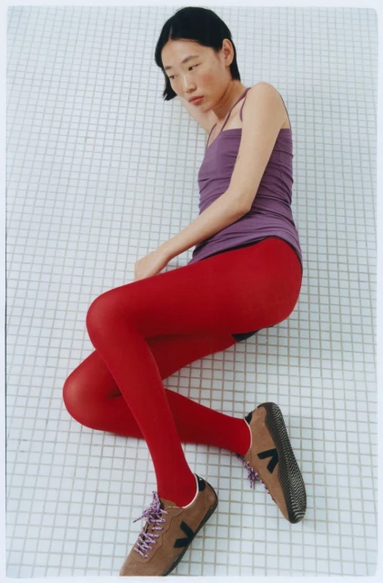 A stylish Asian woman is lying on a white tiled floor, wearing a lilac tank top, red tights, and brown sneakers with a black logo.