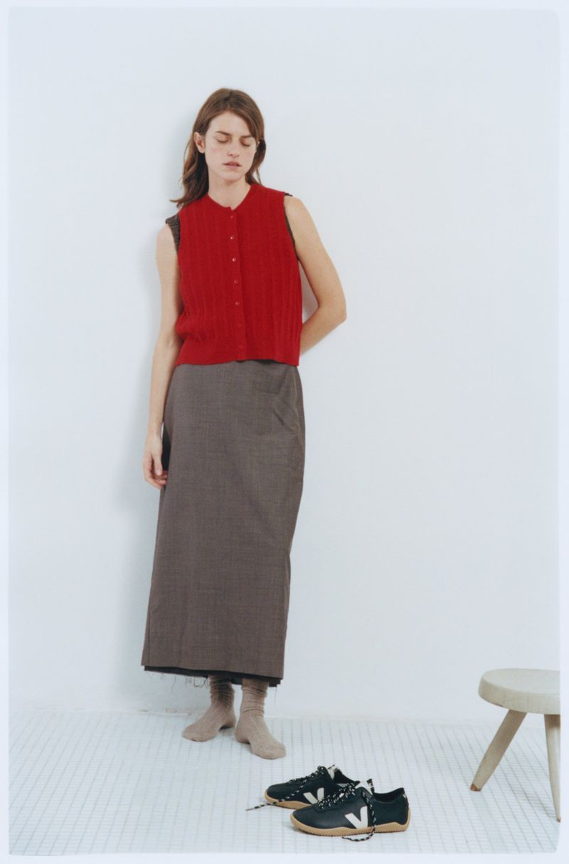 A woman poses indoors wearing a red knit vest, grey skirt, and tan socks, with black sneakers on the floor, against a white wall.