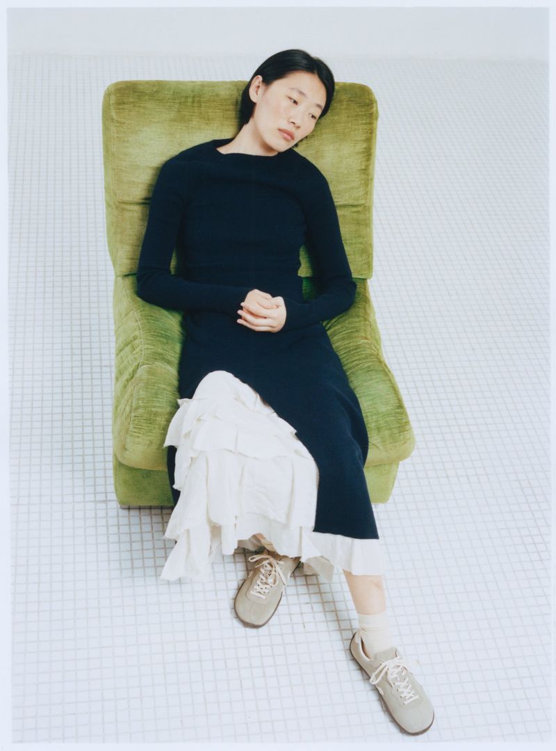 Woman in dark dress and sneakers sits in a green armchair with a relaxed pose on a tile floor in a brightly lit indoor setting.