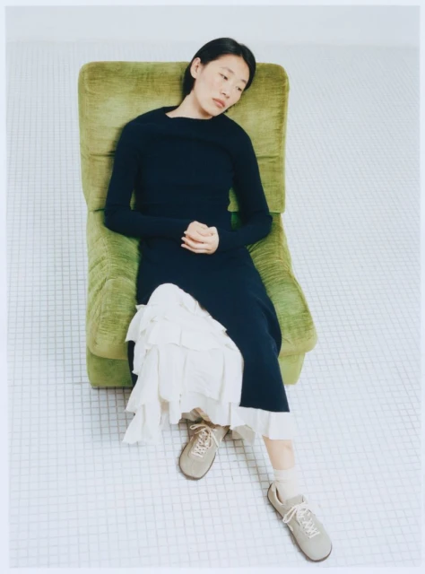 Woman in dark dress and sneakers sits in a green armchair with a relaxed pose on a tile floor in a brightly lit indoor setting.