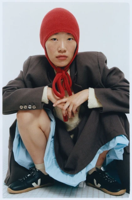 Woman wearing a red bonnet and a grey coat squats while posing indoors, showcasing a unique blend of fashion and comfort.