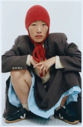 East Asian woman crouching in a red balaclava, oversized dark blazer, and black sneakers against a bright white background.