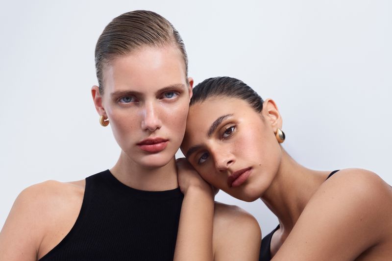 Two adult females pose in a close-up studio shot, highlighting their makeup and minimalist black tops against a clean white backdrop.