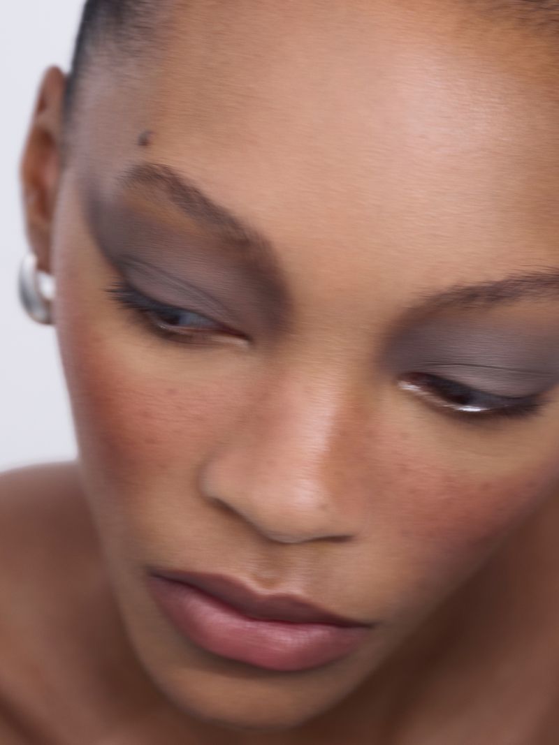 Close-up of a model with dramatic grey eyeshadow and natural makeup, emphasizing facial features and neutral tones against a light backdrop.