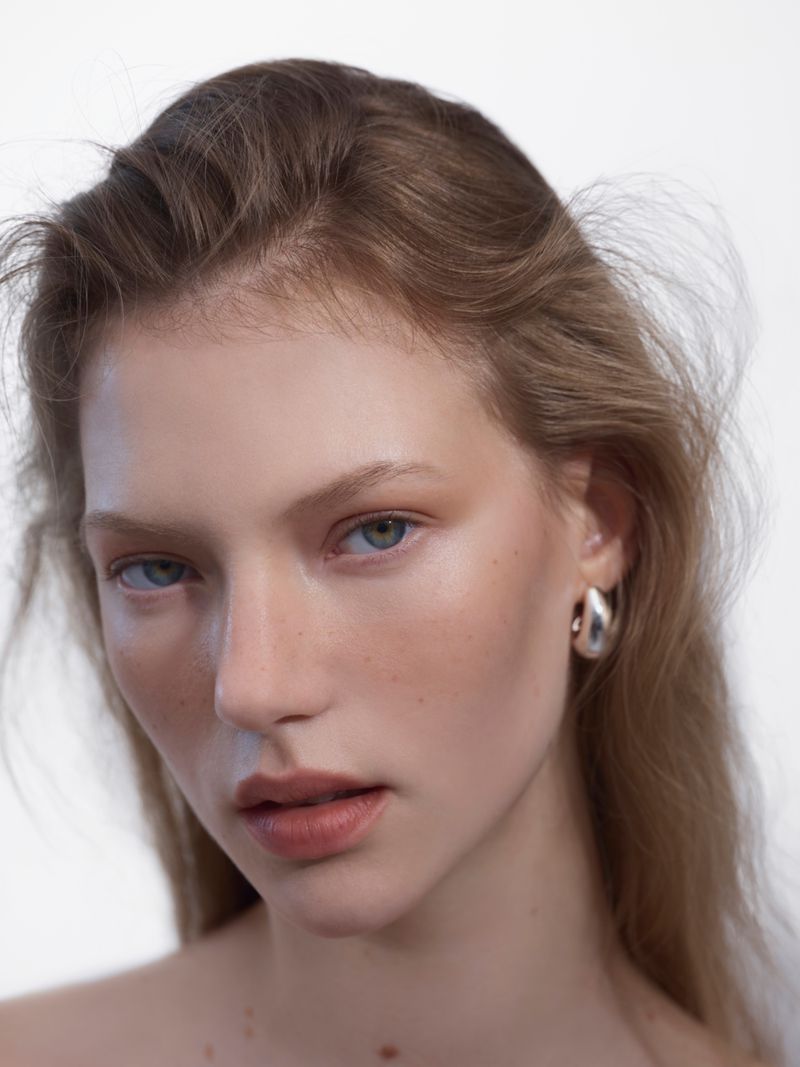 Close-up portrait of a fair-skinned woman with light brown hair, neutral expression, wearing silver hoop earrings against a white backdrop.