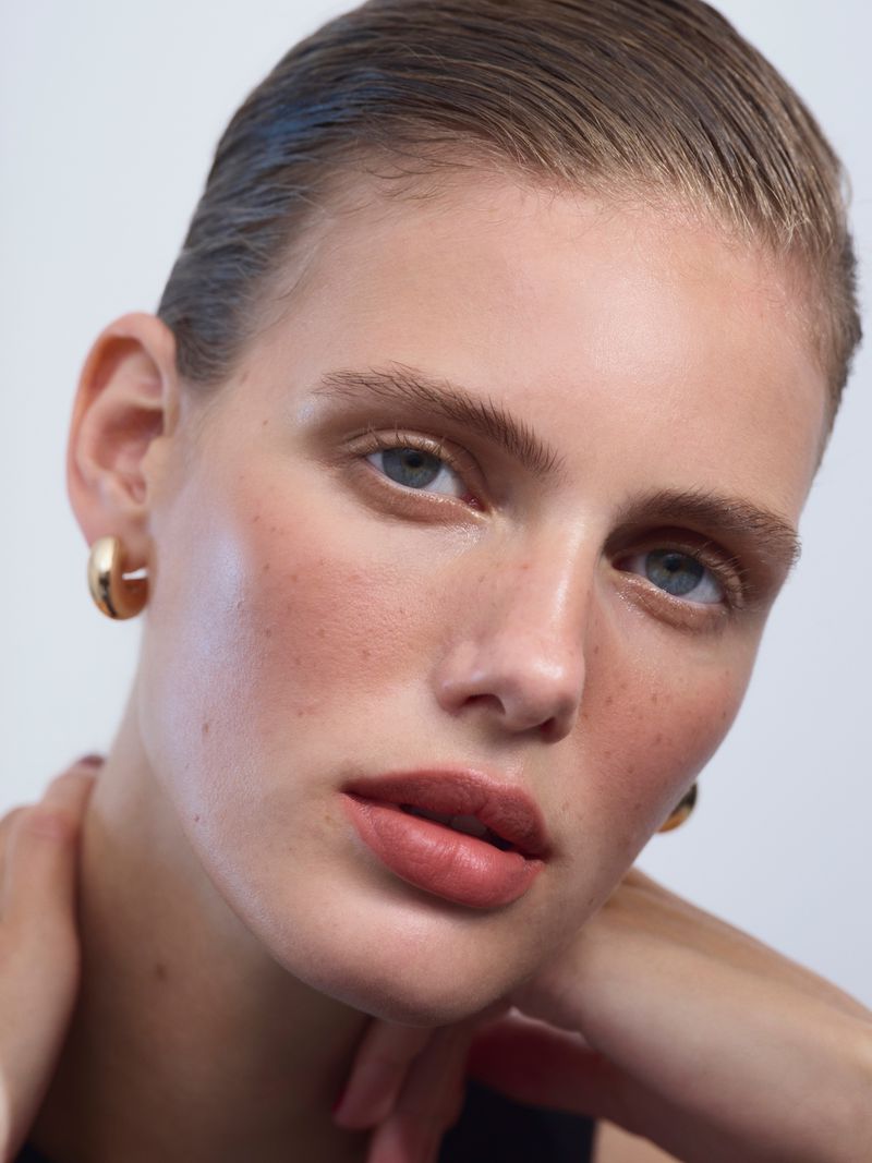 Close-up studio portrait of a young adult woman with slicked-back hair, gold hoop earrings, and bare shoulders against a neutral background.