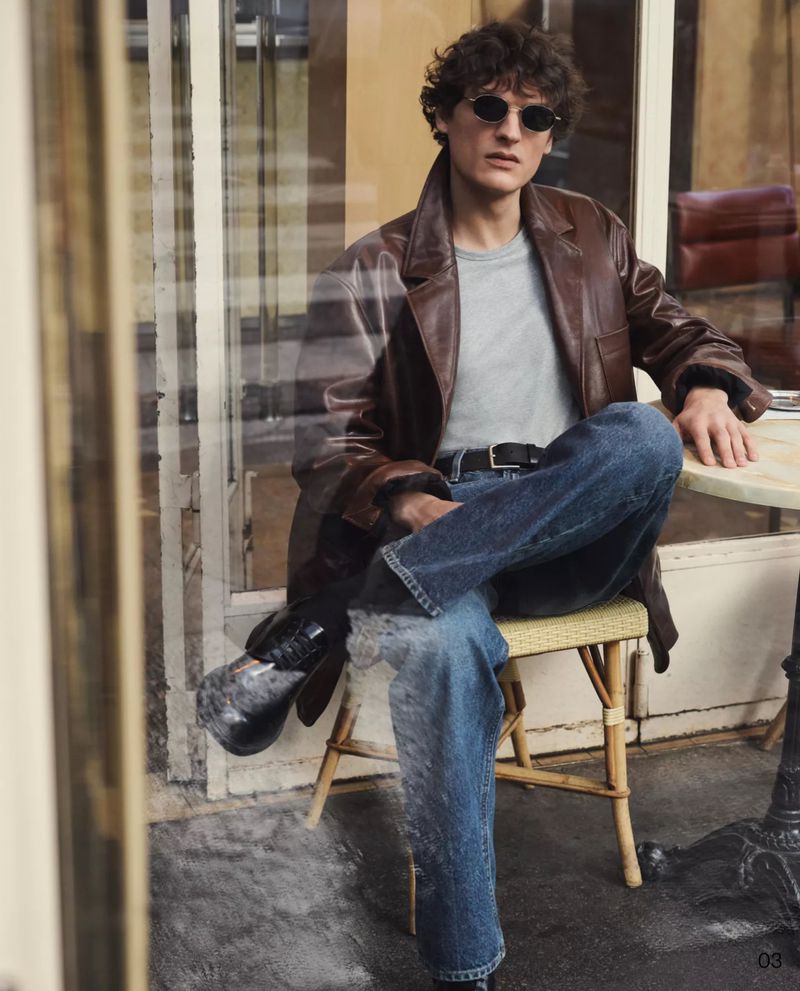 Man in brown jacket and jeans poses while seated at an outdoor cafe with a window reflection in an urban environment.