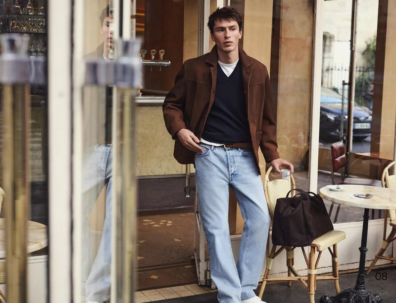 Man in casual fashion stands outside a cafe in daylight wearing a brown jacket, jeans, holding a cup with a brown leather bag on chair.