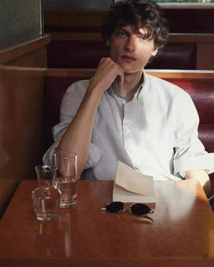 Young male model seated at a wooden table in a diner booth wearing a light patterned button-down shirt.