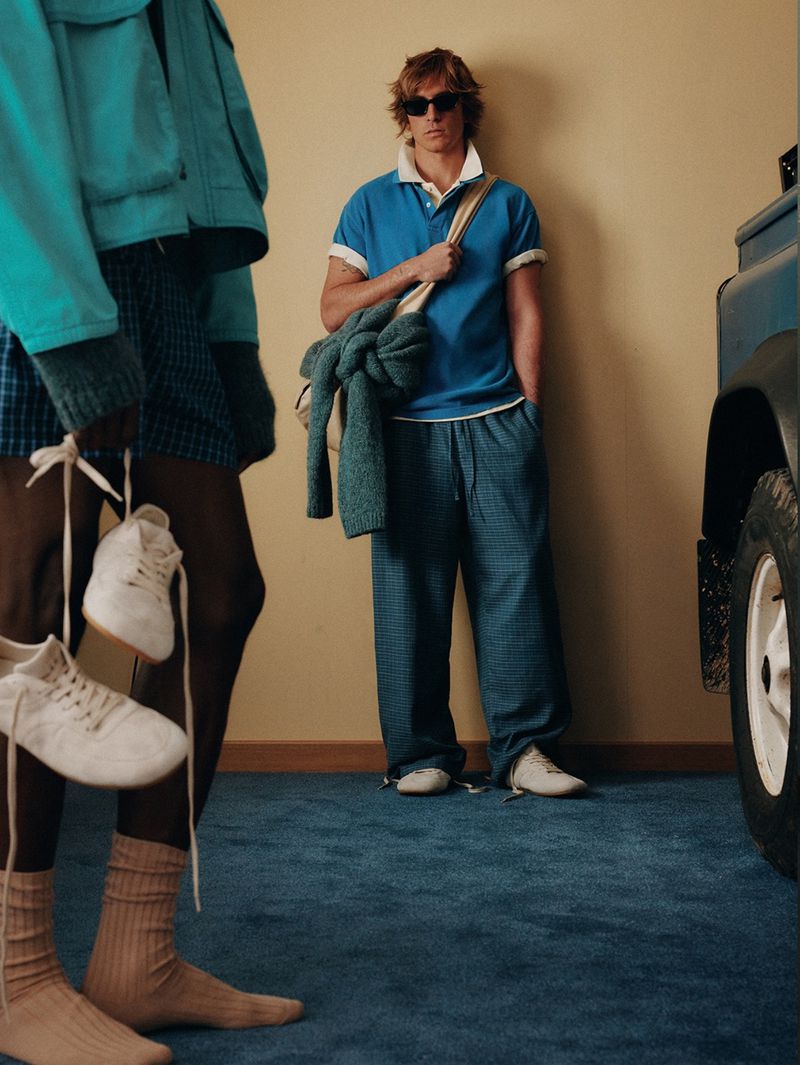 Two adult men are modeling casual outfits, one stands holding sneakers by the laces, and a blue SUV is partially visible in the scene.
