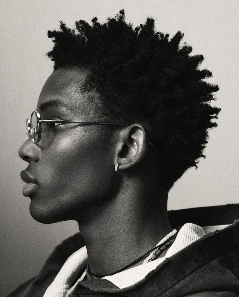 Monochrome portrait of an African-American male in eyeglasses with short afro, wearing a hooded jacket against a neutral backdrop.