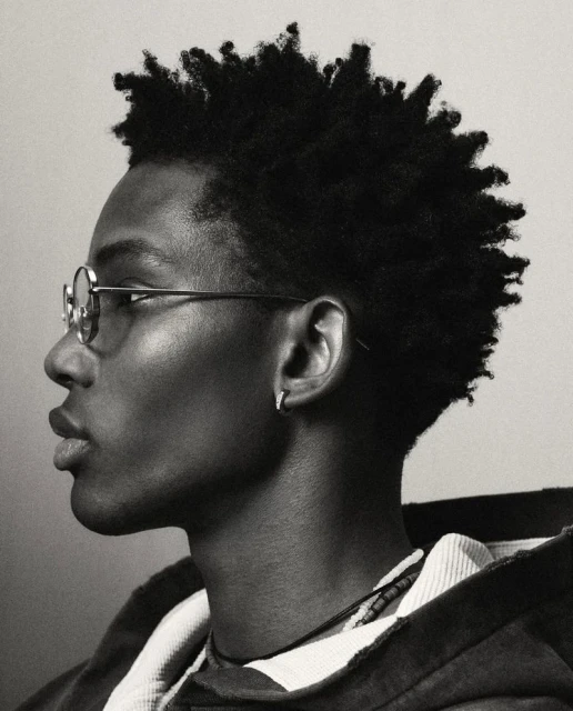 Monochrome portrait of an African-American male in eyeglasses with short afro, wearing a hooded jacket against a neutral backdrop.