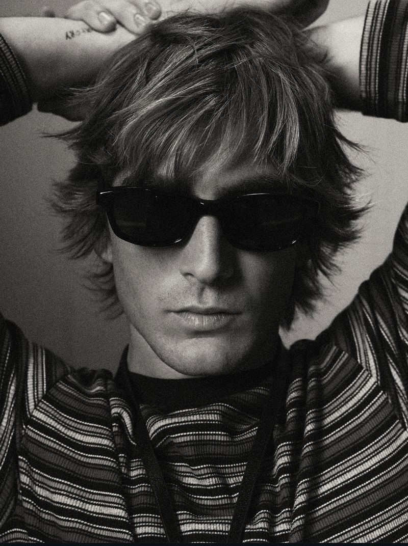 Monochrome portrait of a young adult man with sunglasses and a striped shirt, hands behind his head, with a tattoo on his arm.
