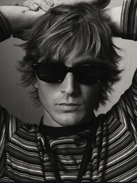 Monochrome portrait of a young adult man with sunglasses and a striped shirt, hands behind his head, with a tattoo on his arm.
