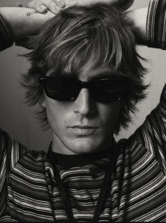 Monochrome portrait of a young adult man with sunglasses and a striped shirt, hands behind his head, with a tattoo on his arm.