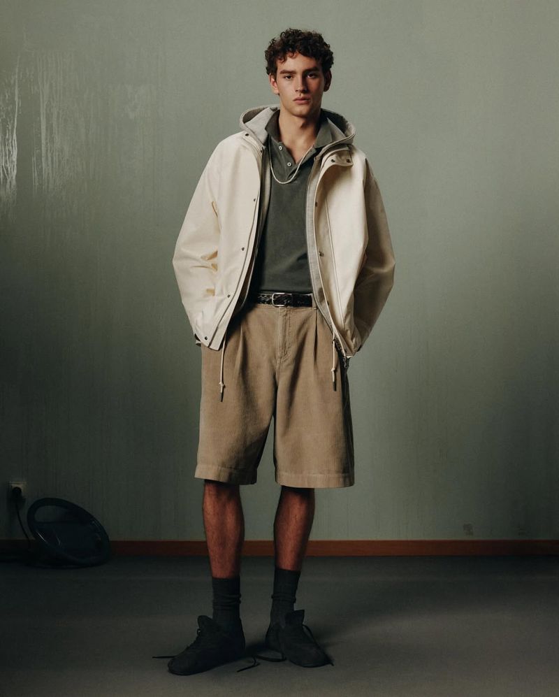 Male model in neutral tones stands in a studio wearing a hooded jacket, polo shirt, belted shorts, socks, and shoes.