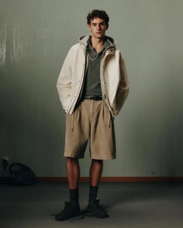 Male model in neutral tones stands in a studio wearing a hooded jacket, polo shirt, belted shorts, socks, and shoes.
