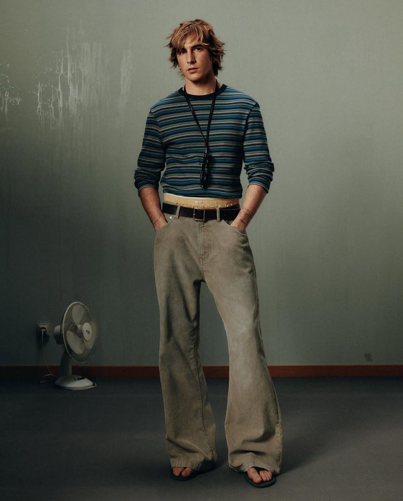 Full-length studio shot of a young adult male with tousled hair, wearing a striped shirt and flared corduroy pants with a retro aesthetic.