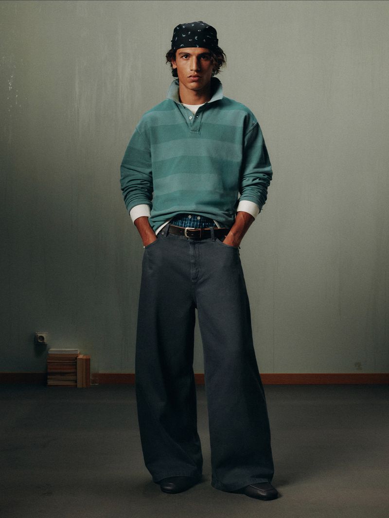 Portrait of a young adult male posing indoors. He is wearing a bandana, striped long-sleeve shirt, baggy pants, and black dress shoes.