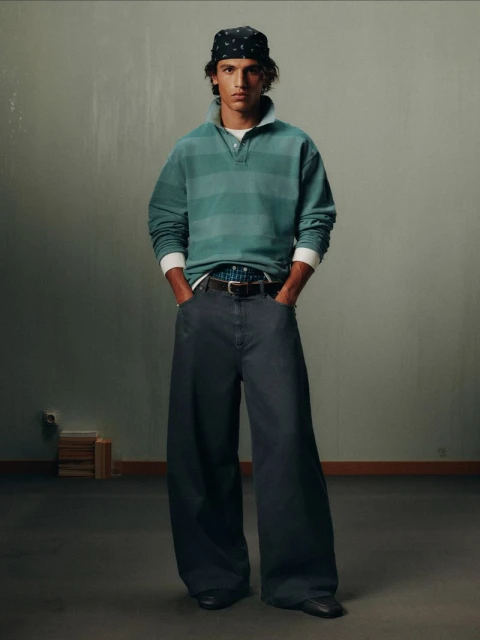 Portrait of a young adult male posing indoors. He is wearing a bandana, striped long-sleeve shirt, baggy pants, and black dress shoes.