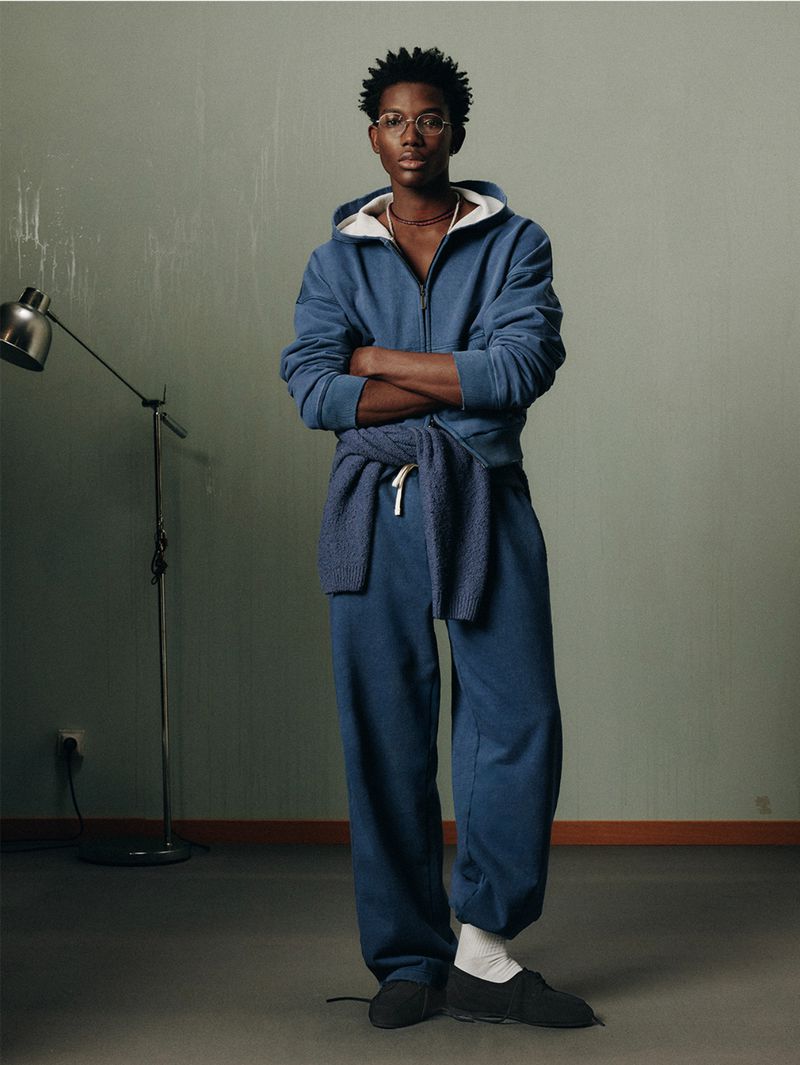 A young Black man in a blue sweatsuit stands indoors with arms crossed, showcasing a casual fashion style against a neutral backdrop.