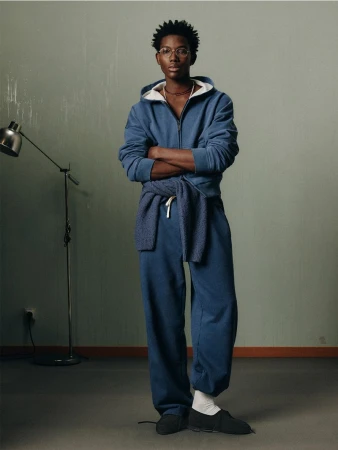 A young Black man in a blue sweatsuit stands indoors with arms crossed, showcasing a casual fashion style against a neutral backdrop.