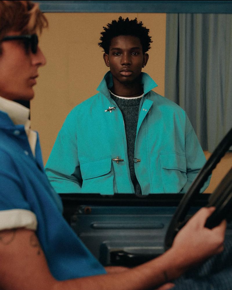 Two adult men in a car, one driving and wearing sunglasses, the other wearing a turquoise jacket, against a neutral background.