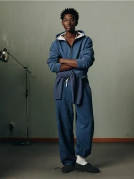 Model standing with arms crossed wearing a blue zip-up hoodie and matching sweatpants against a plain green wall