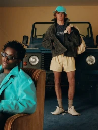 Two male models posing indoors with a vintage blue off-road truck, wearing casual jackets, shorts, cap, and sunglasses.