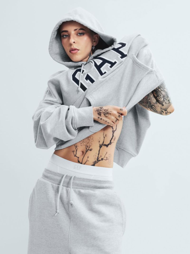Woman in grey GAP hoodie and sweatpants poses in a studio with floral tattoos on her torso and arms, fashion, and lifestyle.