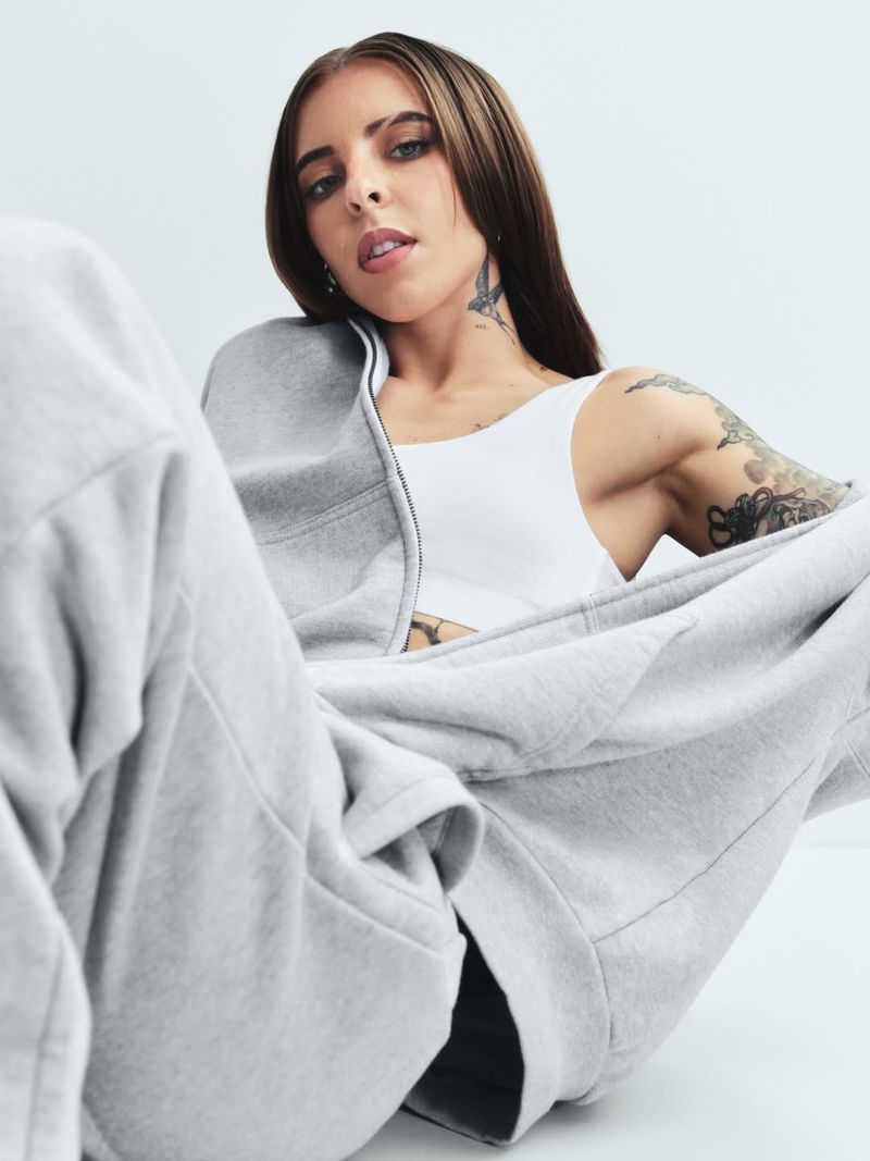 Portrait of a brunette woman in a white tank top and grey sweat suit, posing against a white background in a casual, relaxed manner.