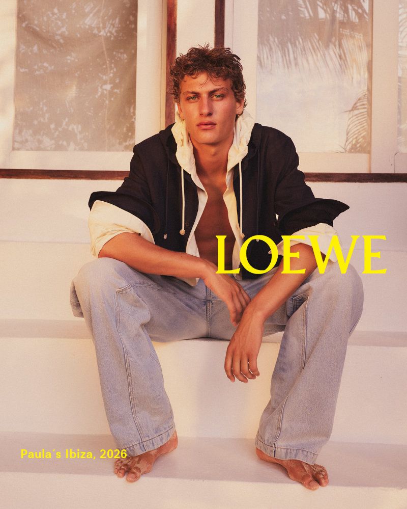 Man models Loewe fashion on stairs. Young male adult sits in jeans and jacket with open shirt, posing against a bright background.