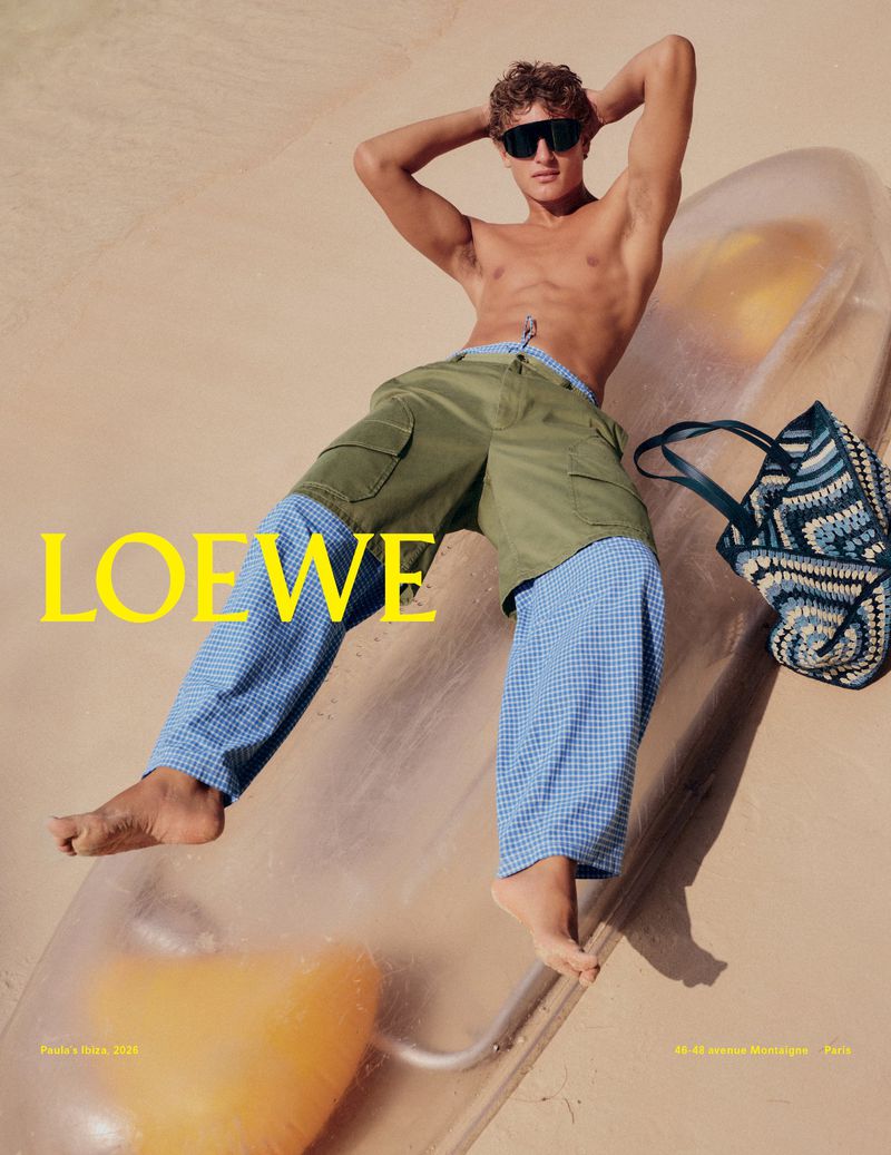 Fashion shot of a male model lying on a transparent inflatable, wearing shorts, with a Loewe beach bag and logo, outdoors.