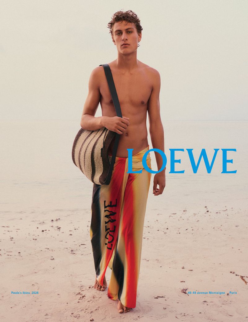 A shirtless male fashion model on a sandy beach carrying a striped tote bag with the Loewe logo visible on the side of his pants.